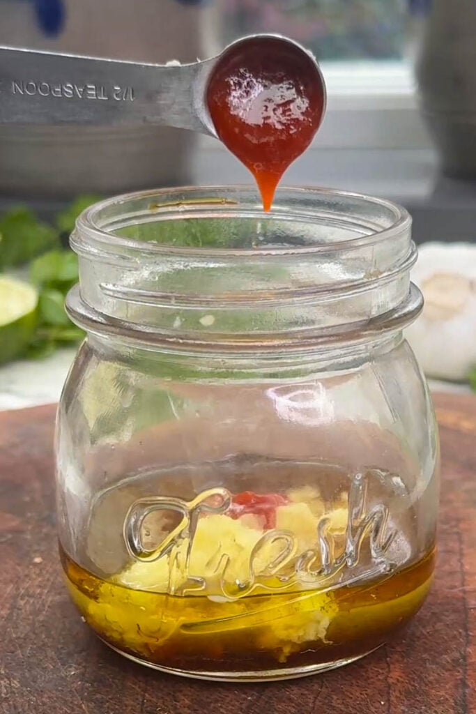 Adding sriracha from a measuring spoon into a glass jar containing oil and minced ginger