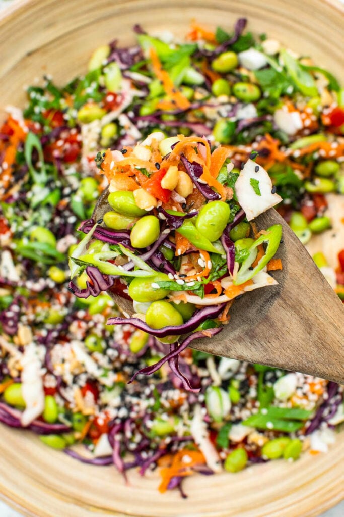 A wooden spoon scoops edamame salad with shredded red cabbage, carrots, peppers, and sesame seeds