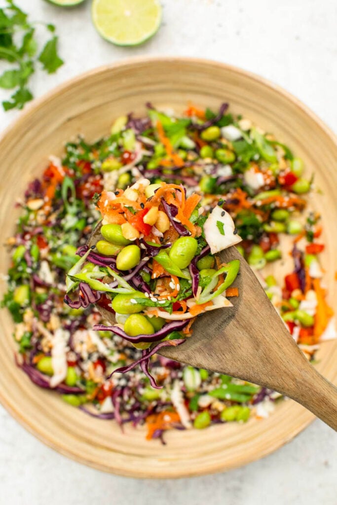 A wooden spoon scoops a serving of vibrant edamame salad with shredded cabbage and carrots from a large bowl