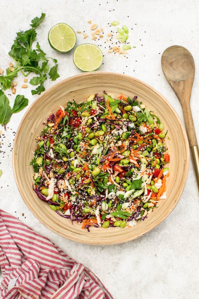 A wooden bowl filled with colorful edamame salad with shredded cabbage, carrots, peppers, herbs, peanuts, and sesame seeds