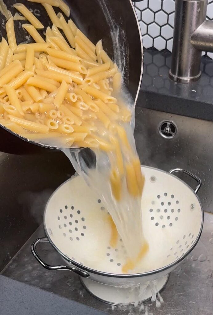 Draining cooked penne in a colander over the sink before adding to puttanesca sauce