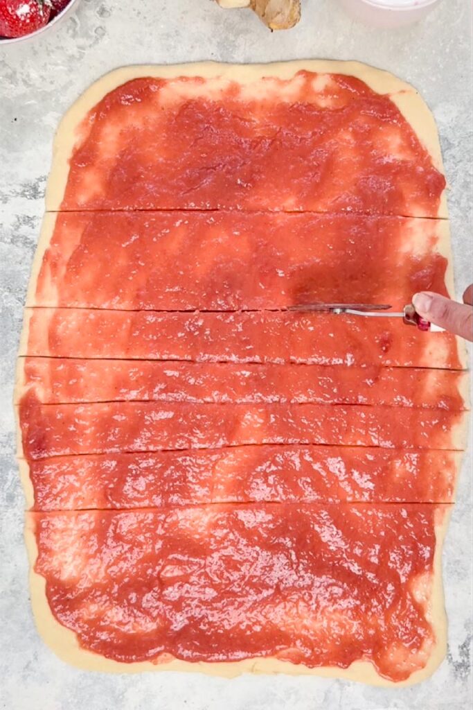 A pizza cutter slicing a rectangle of dough covered in red strawberry rhubarb filling into horizontal strips