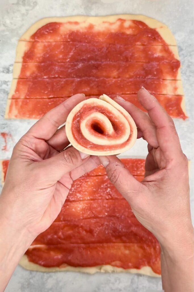 Hands holding a single strip of dough spread with red filling and rolled into a tight swirl