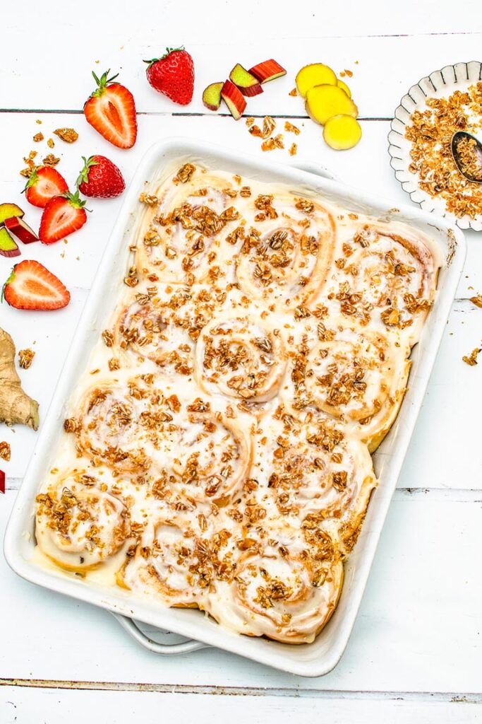 A baking dish of glazed strawberry rhubarb sweet rolls topped with almond praline, surrounded by fresh ingredients
