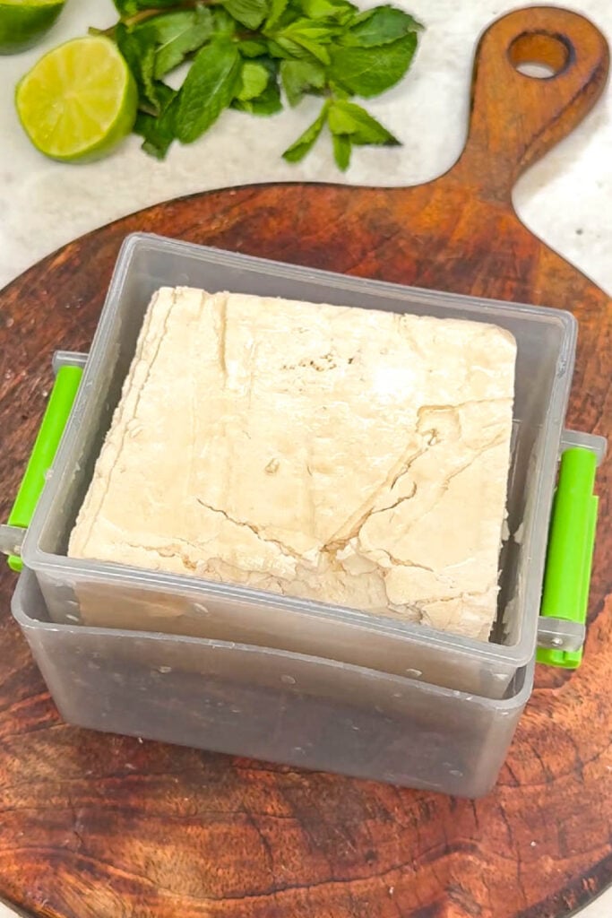 Extra-firm tofu being pressed in a clear plastic tofu press with green handles on a wooden board
