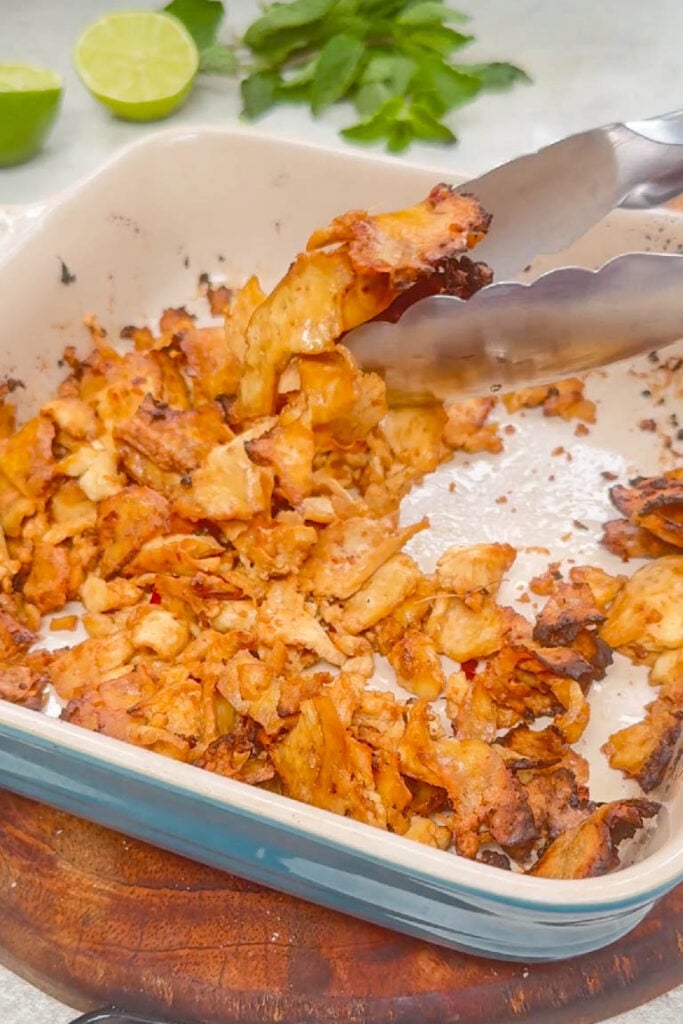 Golden brown, crispy-edged thin tofu strips being lifted with tongs from a square baking dish