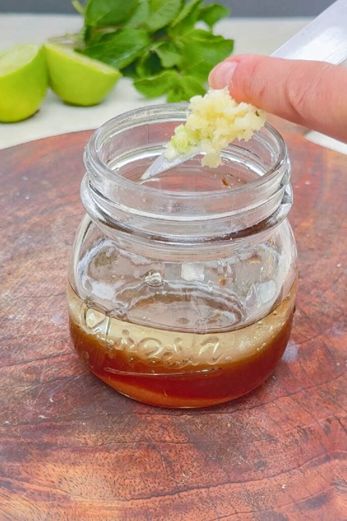 Scraping minced garlic from a knife into a glass jar containing liquid dressing ingredients