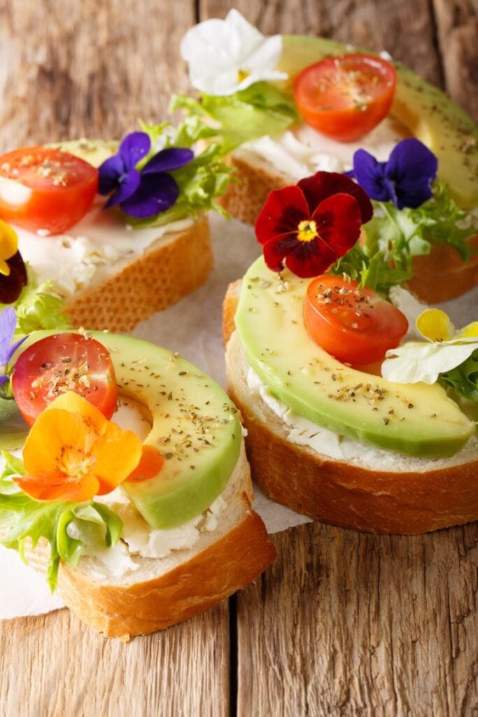 Open-faced sandwiches topped with cream cheese, avocado, cherry tomatoes, and edible flowers on a rustic wooden surface