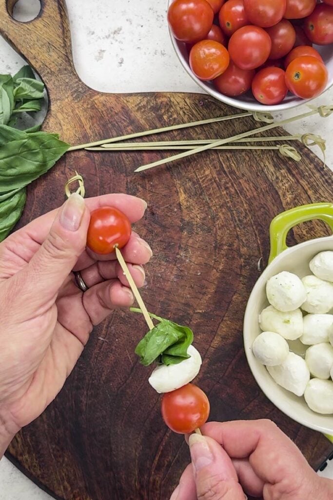 Hands threading a cherry tomato onto a skewer with mozzarella and a folded basil leaf