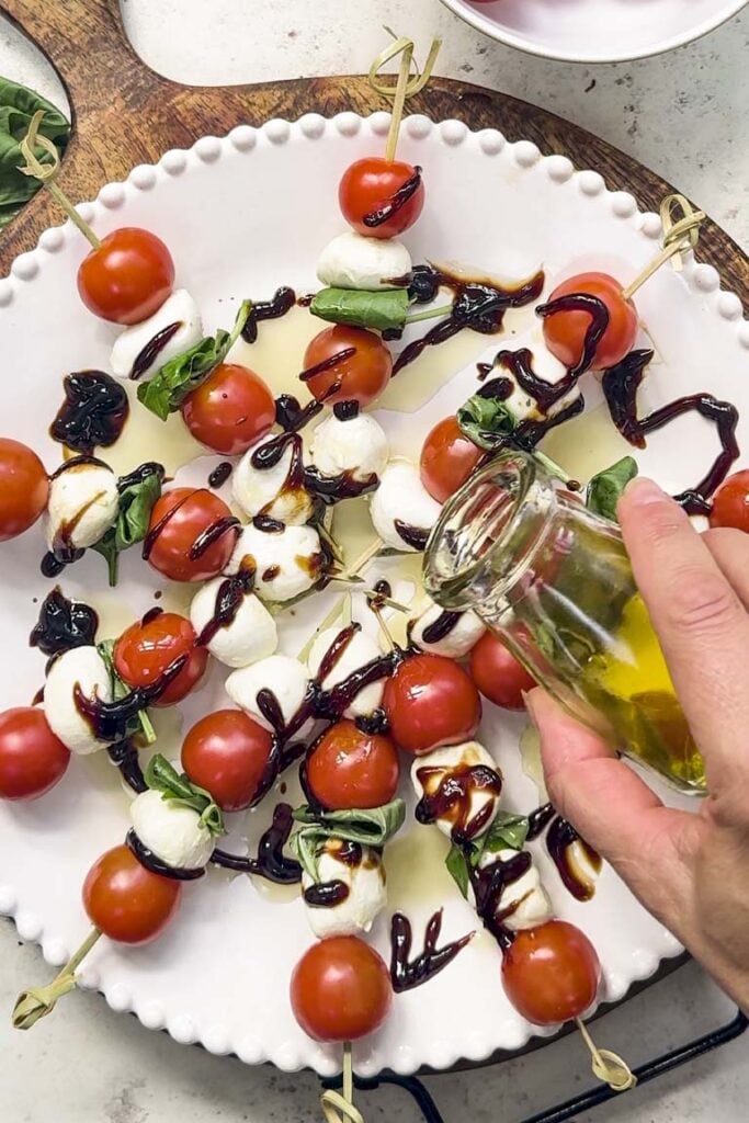 Drizzling olive oil over Caprese skewers with balsamic glaze on a white platter