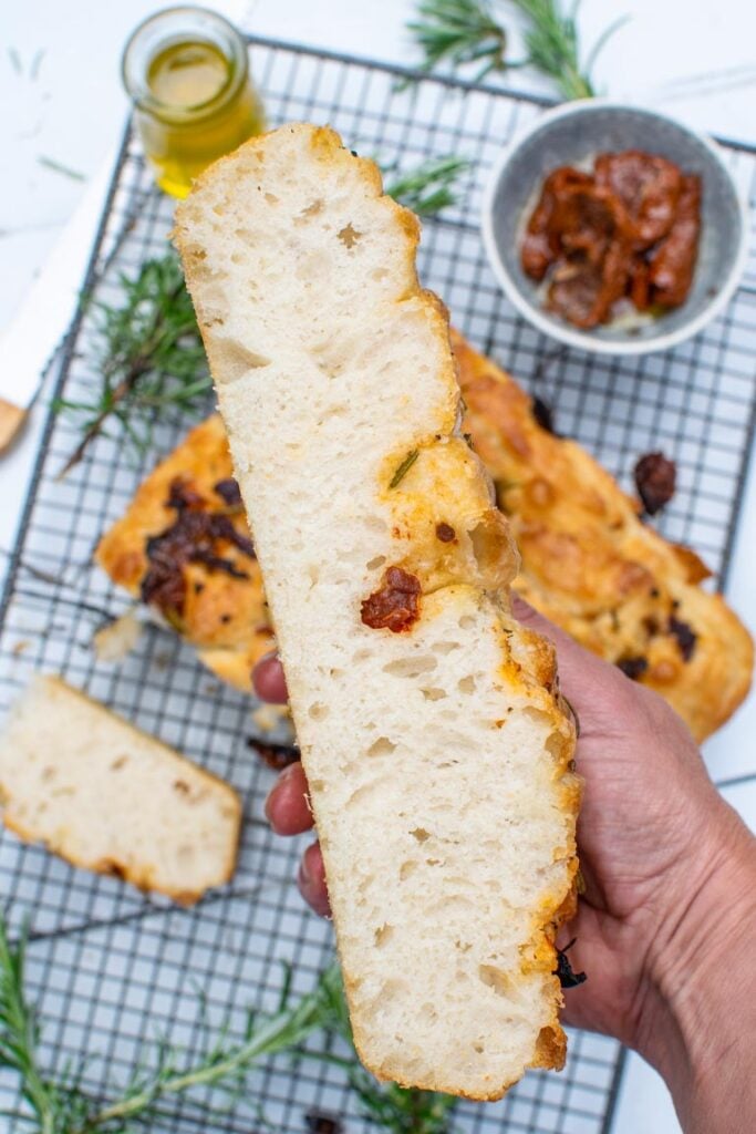 Hand holding a thick slice of focaccia bread showing the airy crumb texture and sun-dried tomato pieces