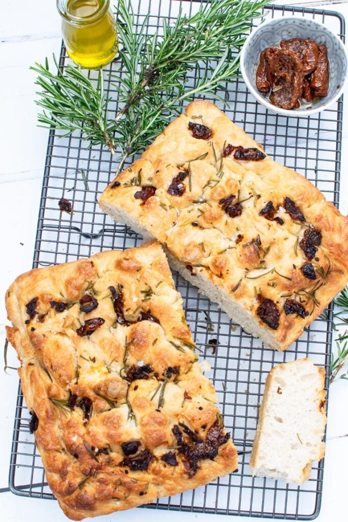 Golden brown focaccia topped with sun-dried tomatoes and rosemary sprigs on a wire cooling rack
