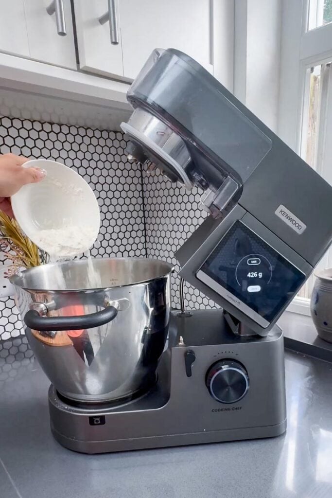 Flour being poured from a white bowl into a stand mixer bowl
