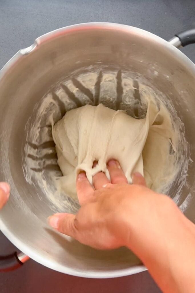 A hand pulls the edge of the dough upward and folds it toward the center of a metal mixing bowl