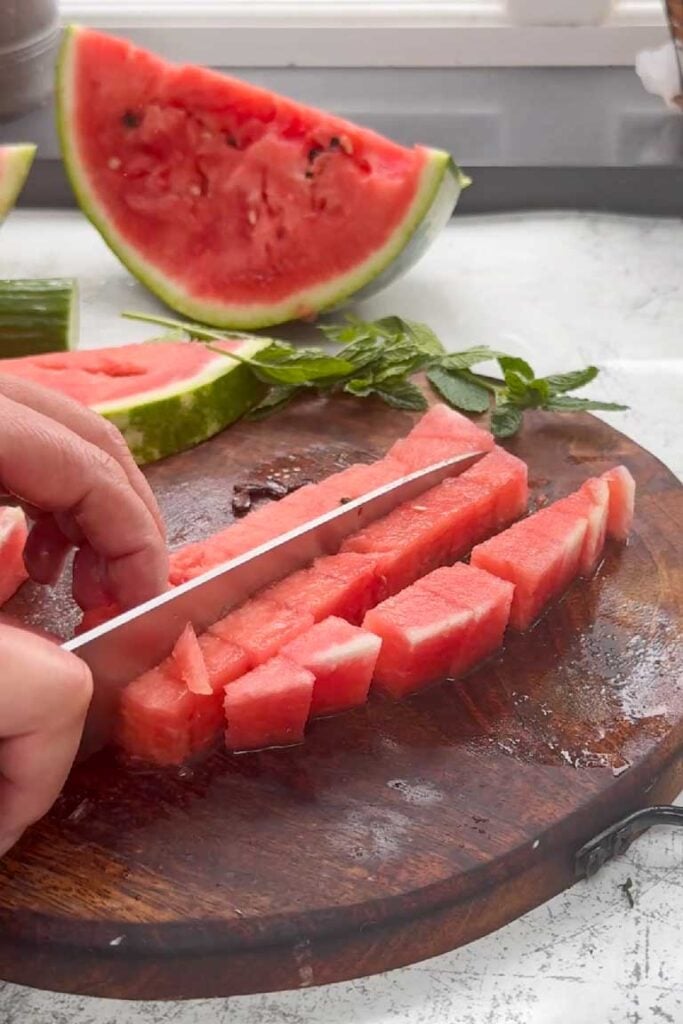 Hands dicing fresh watermelon into cubes on a wooden chopping board