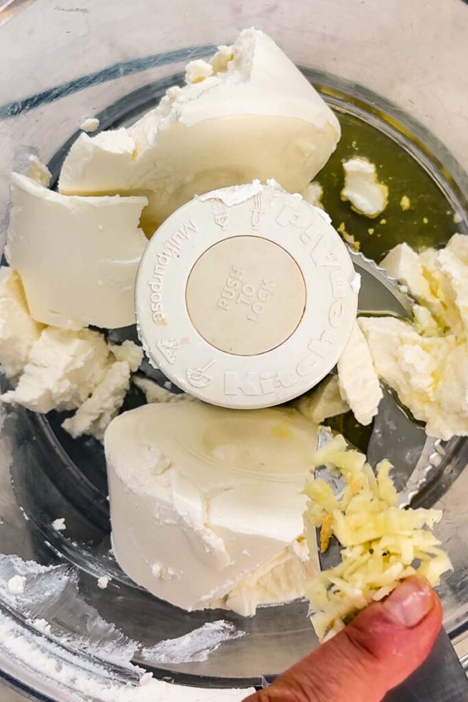 Ricotta cheese and olive oil in a food processor bowl with minced garlic being added