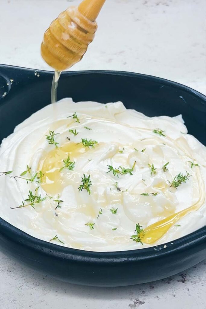 Smooth whipped ricotta in a black bowl, topped with fresh thyme and a drizzle of honey from a wooden dipper