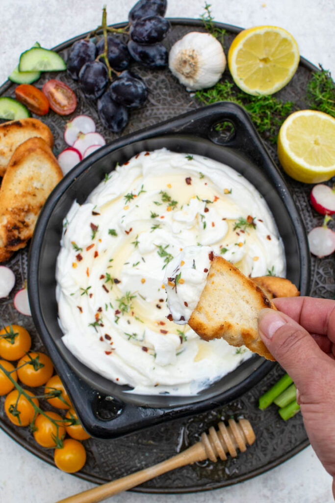 A hand dipping toasted bread into a bowl of whipped feta topped with honey, herbs, and red pepper flakes