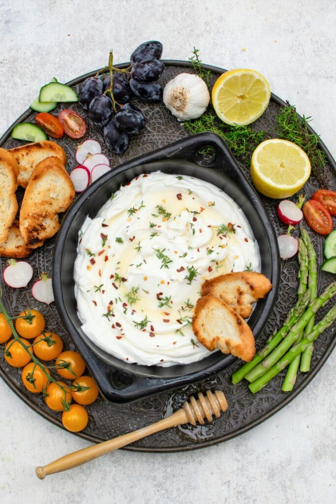 Whipped feta dip topped with honey and herbs, served on a platter with crostini and fresh vegetables