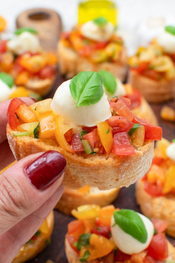 Hand holding a slice of Caprese Bruschetta topped with tomatoes and mozzarella