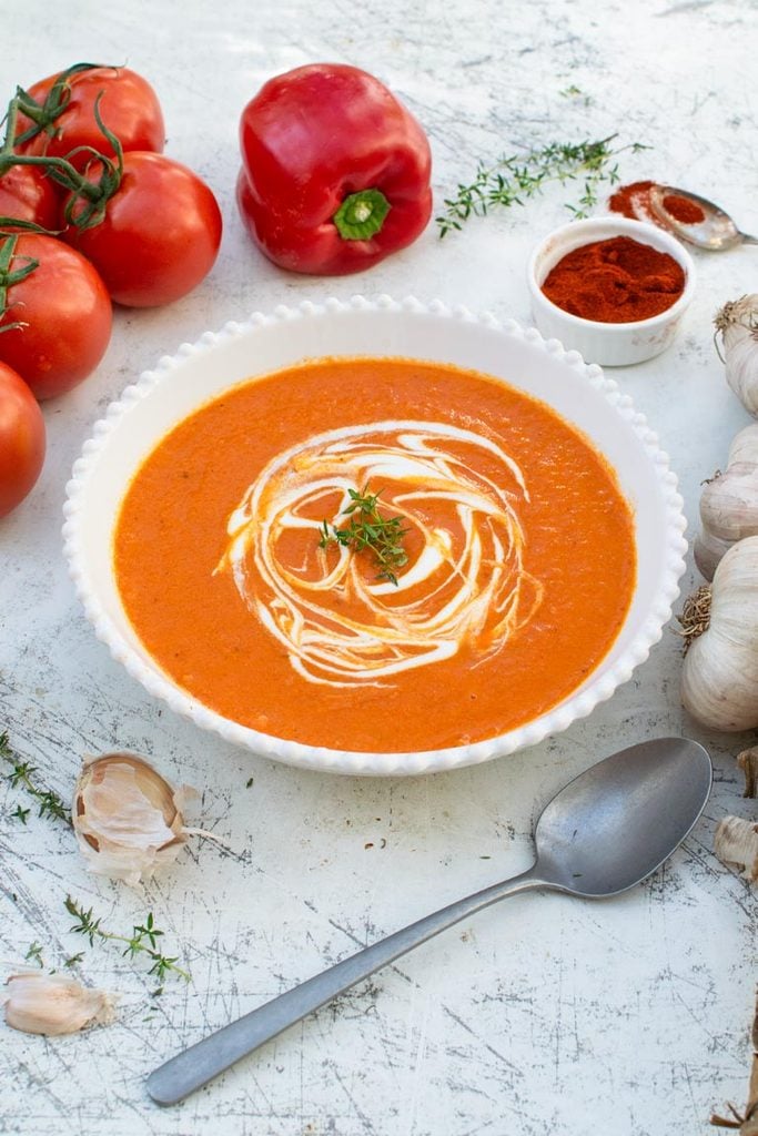 Side shot shot of roasted red pepper and tomato soup in a white bowl with spoon on the side
