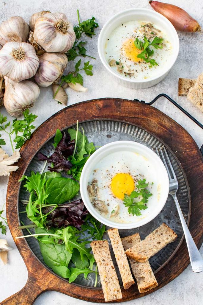 Oeufs en cocotte with mushrooms served on a board with bread soldiers, salad, garlic and shallots