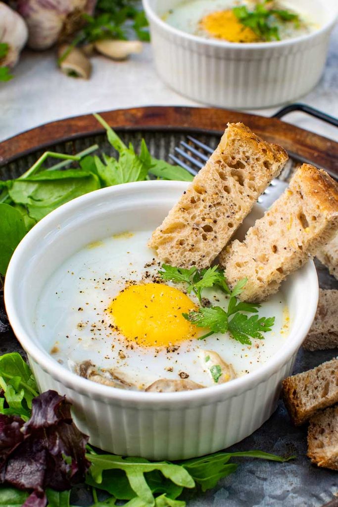 Close-up of baked eggs with mushrooms topped with parsley and black pepper, served with sourdough soldiers