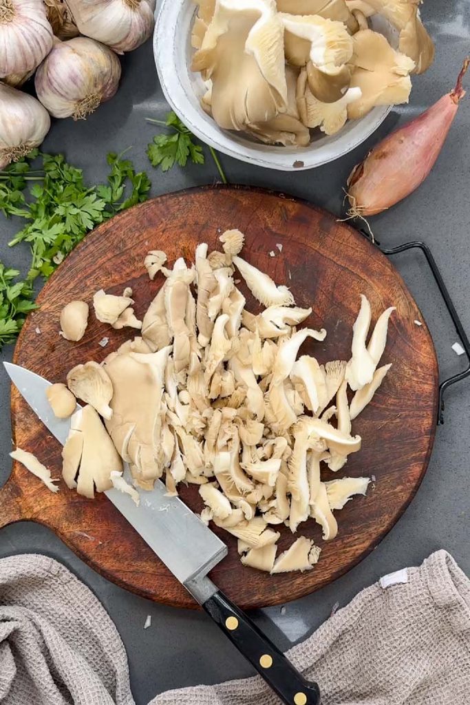 Sliced oyster mushrooms on a wooden chopping board with a knife, shallot, garlic and parsley
