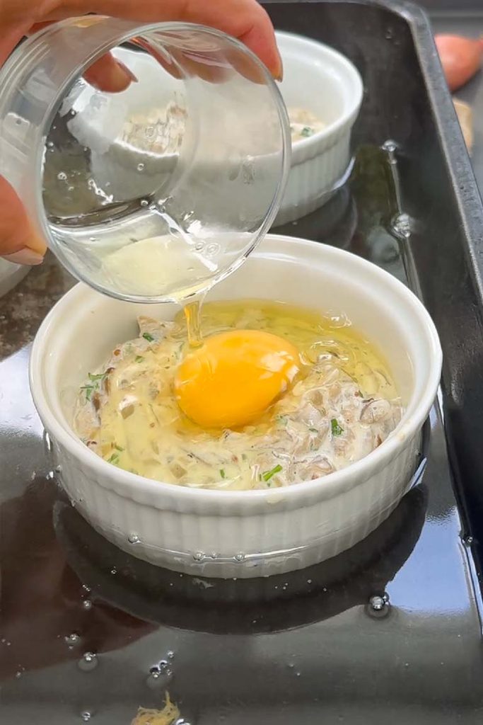 Egg being poured into a ramekin of creamy mushrooms set in a roasting tin for a water bath