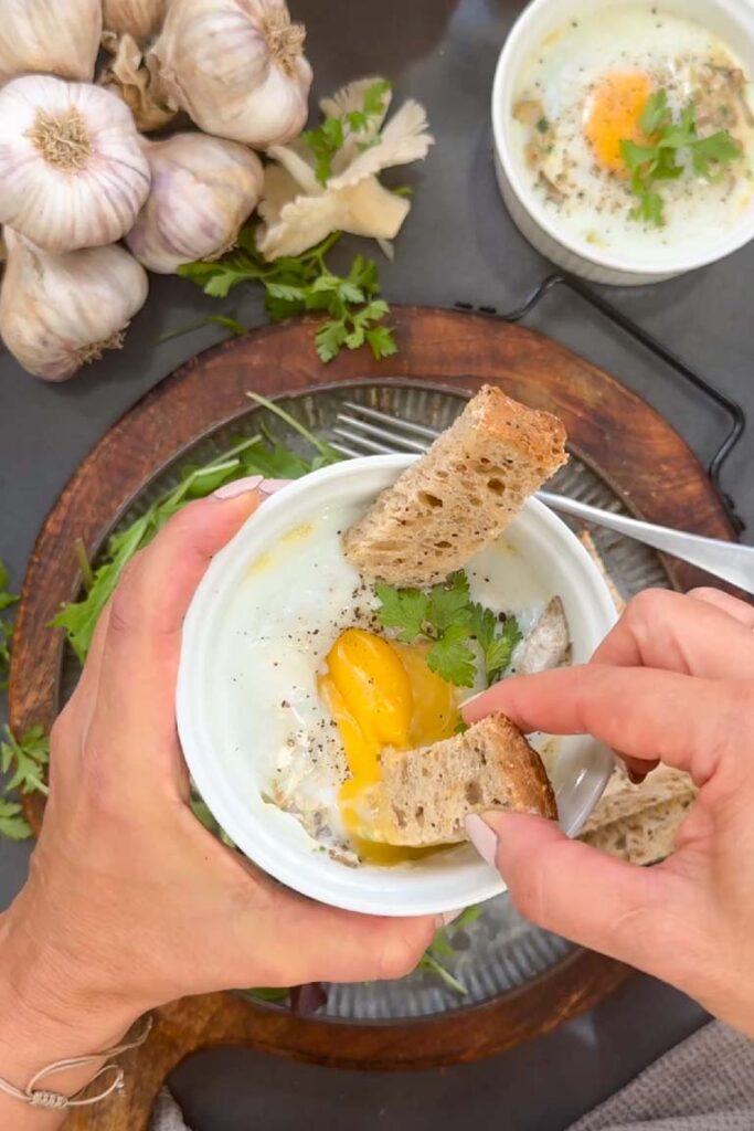 Hand dipping a bread soldier into a baked egg with mushrooms