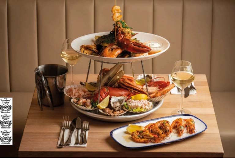 Seafood platter tower with lobster, prawns and oysters beside a plate of grilled prawns and glasses of white wine at a London restaurant