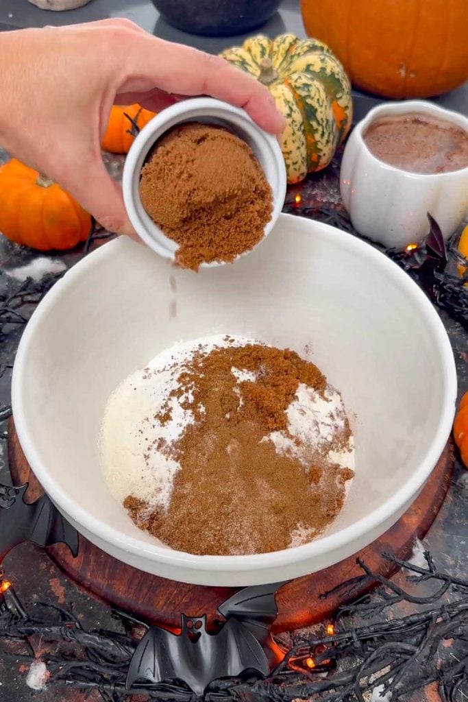 Mixed spice being added to a bowl of flour, baking powder and dark brown sugar for pumpkin pancake batter