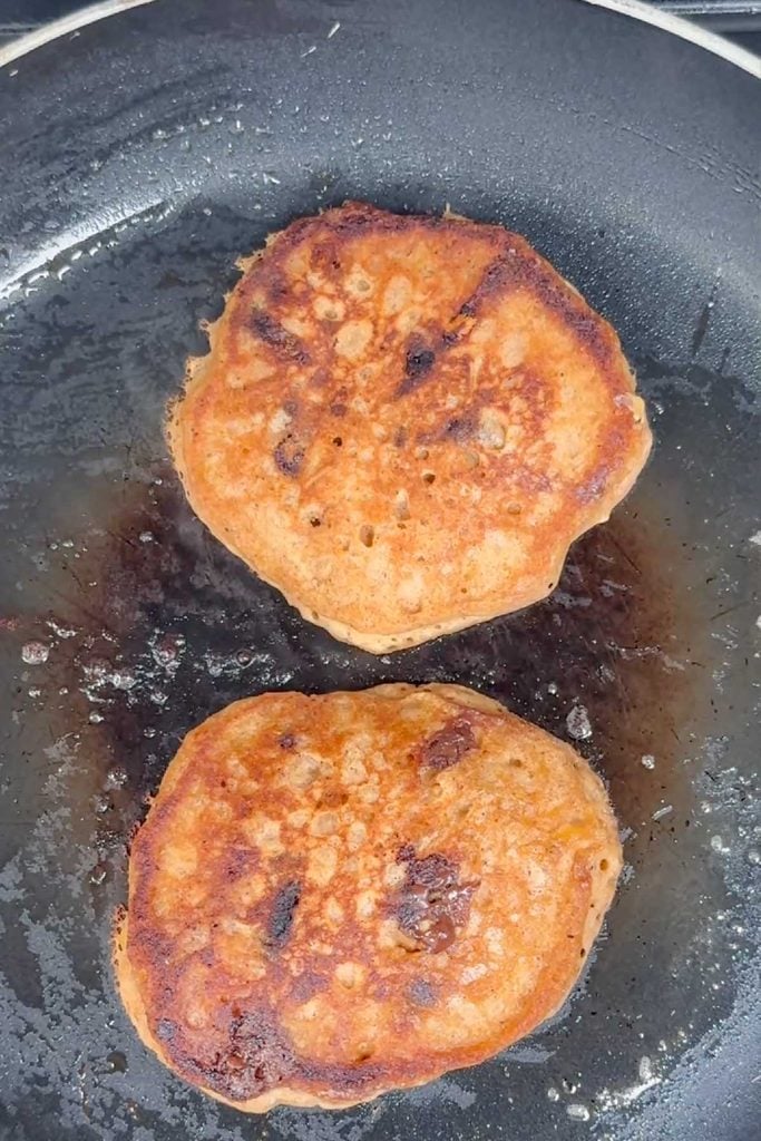 Two golden pumpkin pancakes cooking in a frying pan