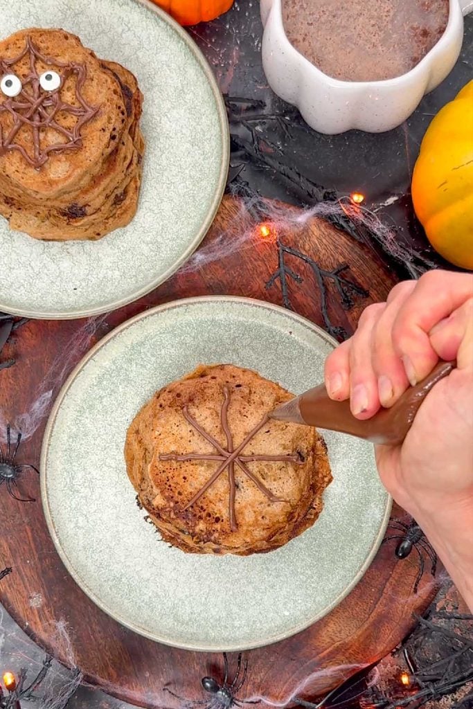 Piping melted chocolate lines from the centre outwards on a stack of Halloween pancakes to start a spider web