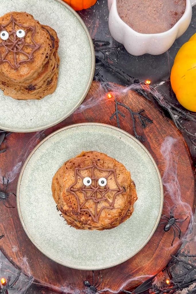 Stack of pumpkin pancakes with a chocolate spider web and two edible googly eyes on a grey plate