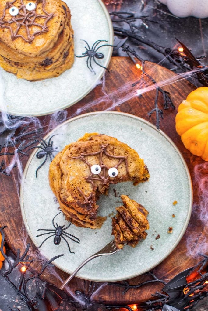 Overhead view of a stack of Halloween pancakes with a bite taken out, surrounded by plastic spiders, cobwebs and mini pumpkins