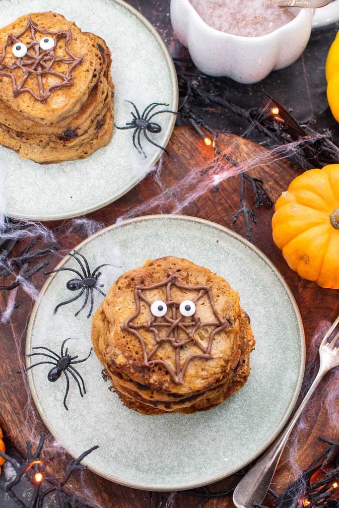 Overhead shot of two stacks of pumpkin Halloween pancakes with a chocolate spider web and edible eyes, surrounded by plastic spiders, cobwebs and mini pumpkins