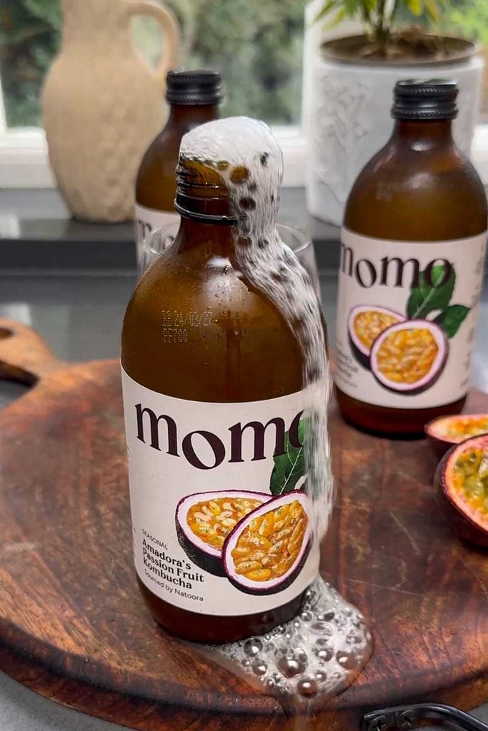 Three bottles of MOMO passion fruit kombucha with halved passion fruit and green plants behind