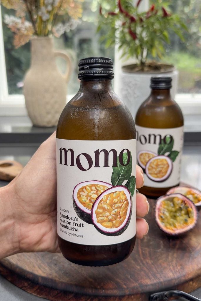 Hand holding a bottle of MOMO x Natoora passion fruit kombucha beside a halved passion fruit
