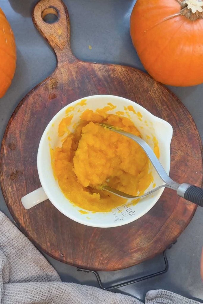 Steamed pumpkin mashed to a smooth puree in a white jug with a potato masher, for pumpkin shaped bread