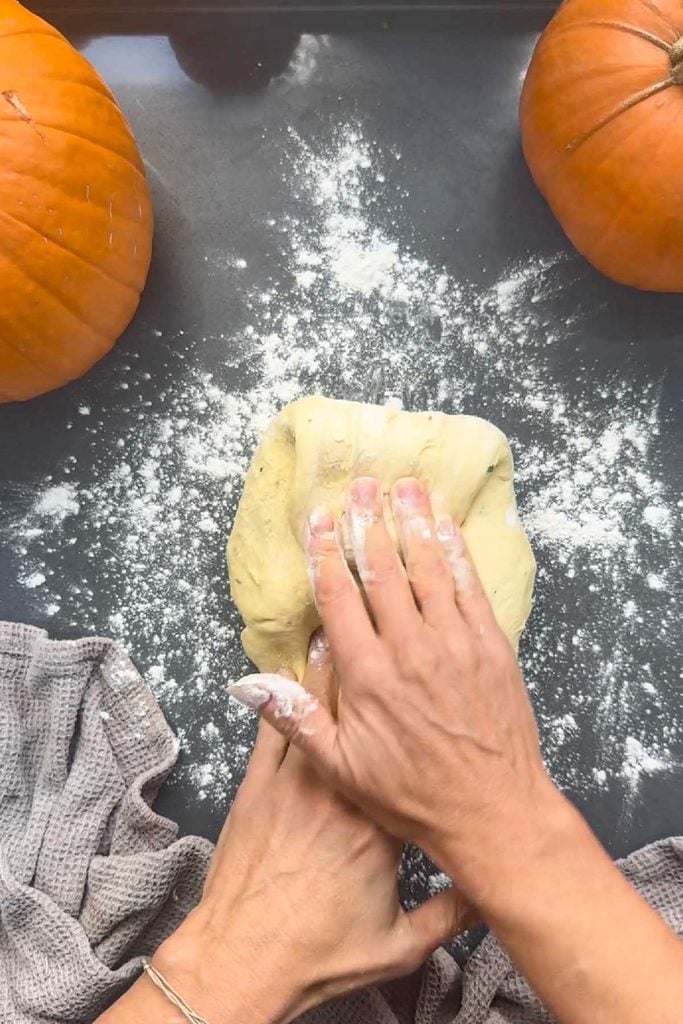 Hands stretching and folding pumpkin bread dough on a floured worktop to shape it into a round
