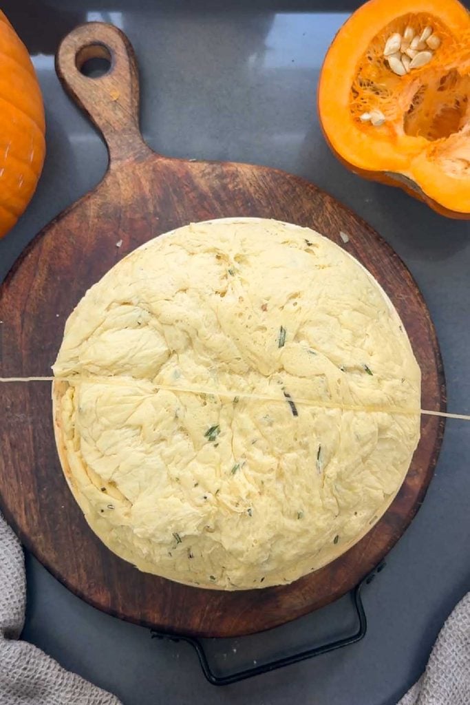 Round of pumpkin and rosemary dough on a wooden board with the first piece of oiled kitchen twine laid across it