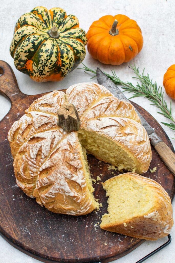 Pumpkin shaped bread cut open on a wooden board to show the soft rosemary crumb, with a knife and fresh rosemary