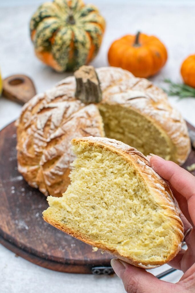 Hand holding a wedge of rosemary pumpkin bread showing the soft yellow crumb, with the rest of the loaf behind