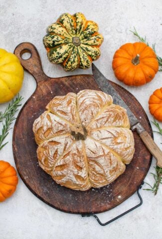 Pumpkin shaped bread with a scored, floured crust on a wooden board, surrounded by mini pumpkins, squash and fresh rosemary