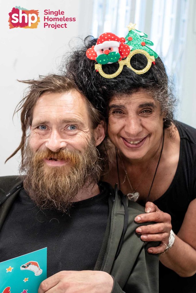 Smiling man and woman in novelty Christmas glasses holding a Christmas card at a Single Homeless Project celebration