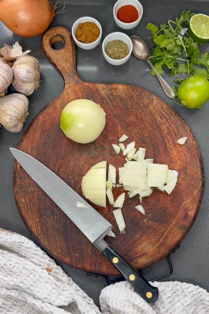 Diced onion on a wooden chopping board with garlic, lime, coriander and small bowls of spices