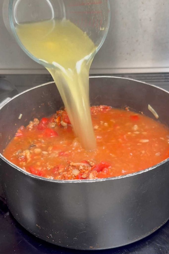 Vegetable stock being poured into the tomato and red pepper base of taco soup
