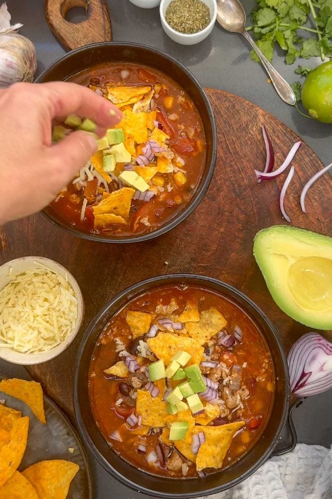 Topping a bowl of taco soup with tortilla chips, with avocado, grated cheese and red onion on the side