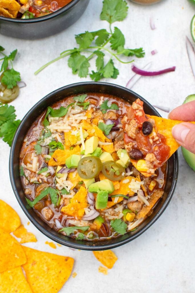 Tortilla chip being dipped into a bowl of vegetarian taco soup topped with avocado, cheese and jalapeños
